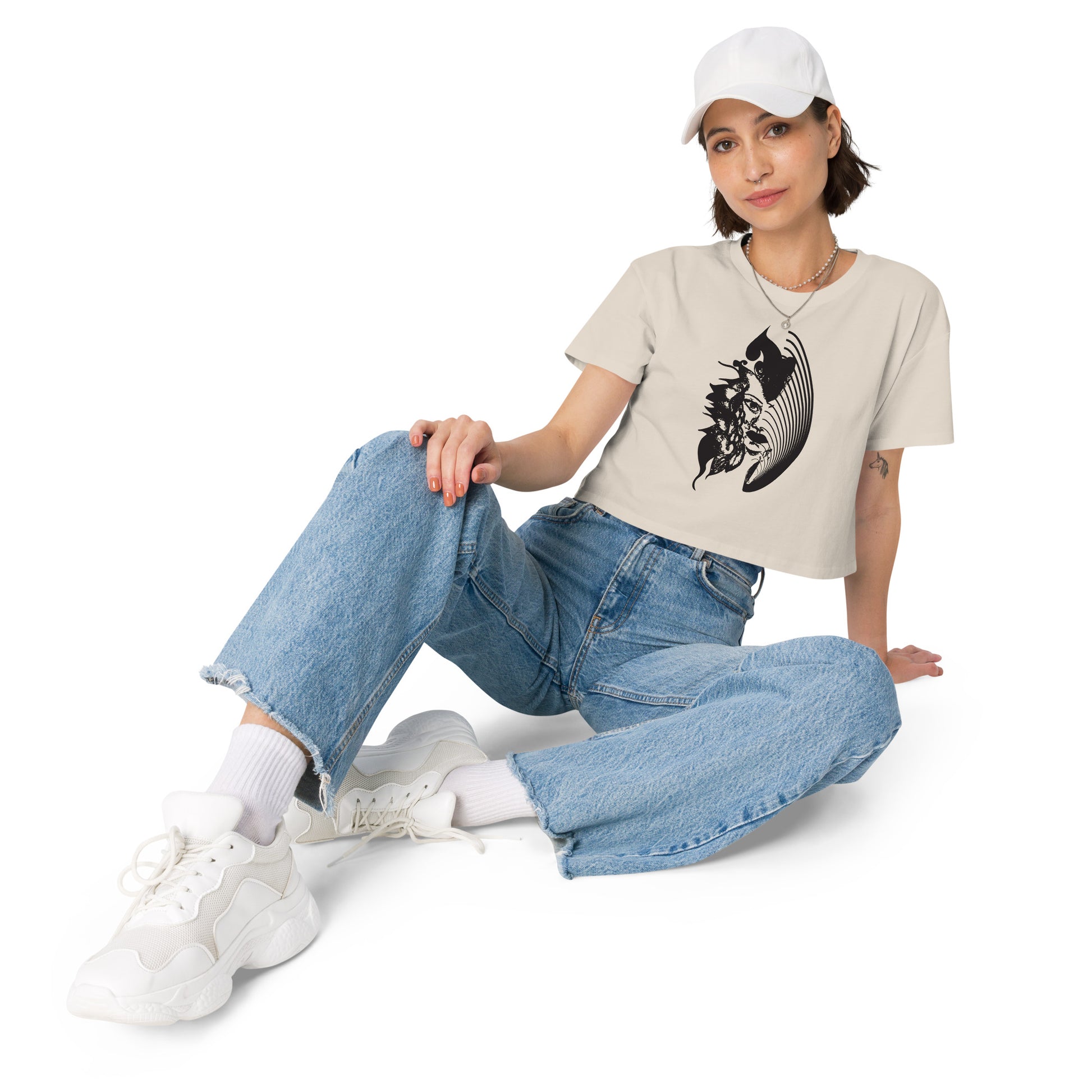 A model wearing a bone K.L.O.E. crop t-shirt featuring a culturally inspired, hand-drawn black graphic of a bold, expressive face surrounded by intricate details, celebrating individuality and artistic self-expression in modern bohemian style fashion.