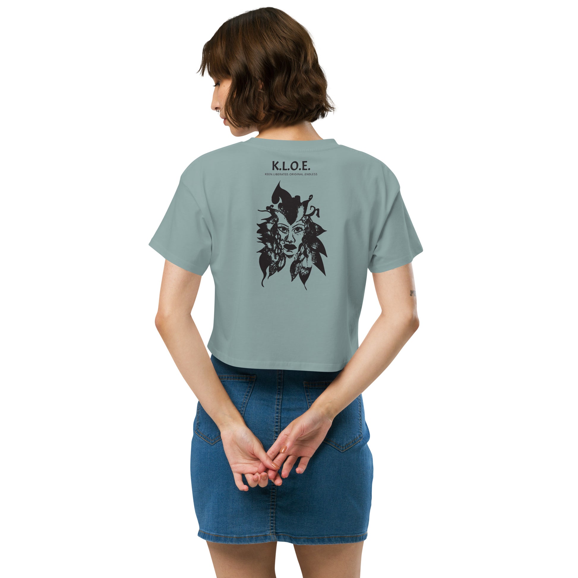 Back view of a mineral K.L.O.E. crop t-shirt showcasing a culturally inspired, hand-drawn black design featuring a bold, expressive face intertwined with flowing, abstract patterns, and K.L.O.E. logo, embodying artistic self-expression and modern bohemian style fashion, on a model.